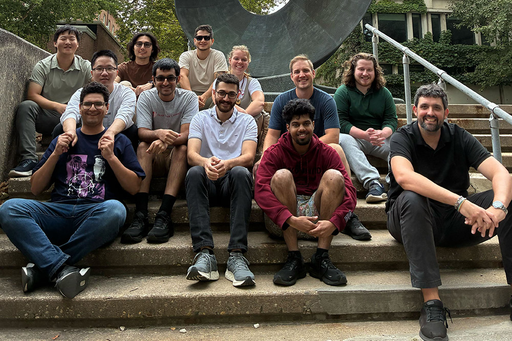 Jesus Perez Rios and Research team Group of eleven individuals From The Stony Brook University Department of Physics and Astronomy posing on steps outdoors, smiling, with a large sculpture in the background.