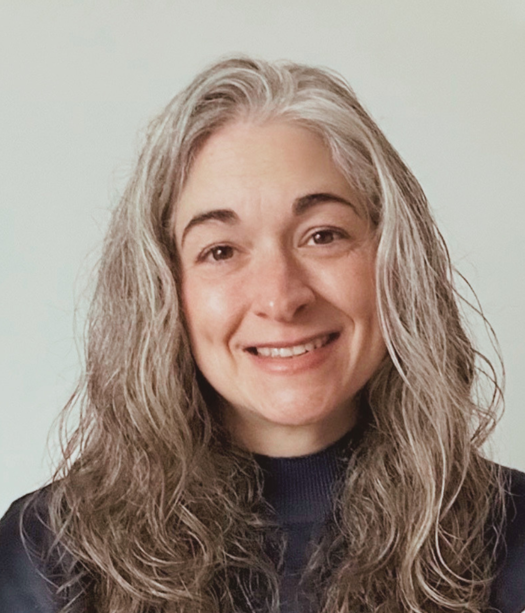 Smiling woman with long, wavy gray hair against a plain backdrop. She wears a dark top and exudes warmth and approachability.