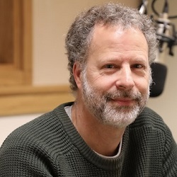 Man with facial hair, wearing a sweater, sits in front of a large microphone