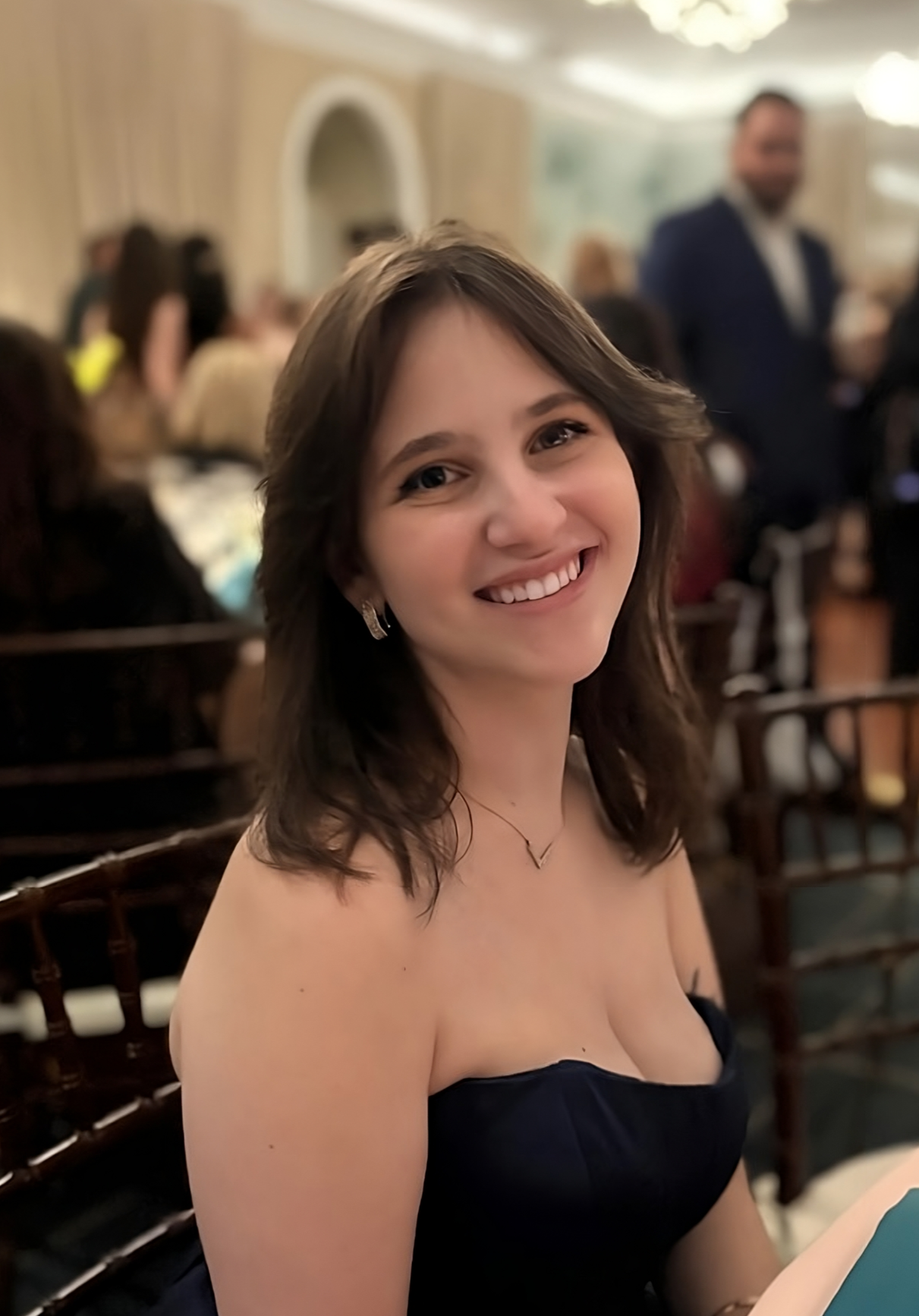 a smiling person wearing a formal dress sits on a chair in a banquet hall 
