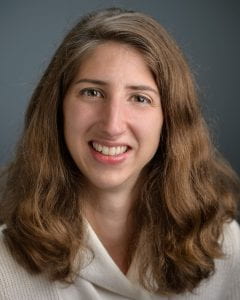 Portrait of a smiling person with long, wavy hair, wearing a light-colored scarf over a blouse against a solid dark background.