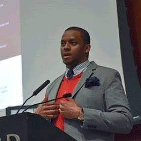 A person speaking at a lectern with a microphone, wearing a gray suit and red sweater.