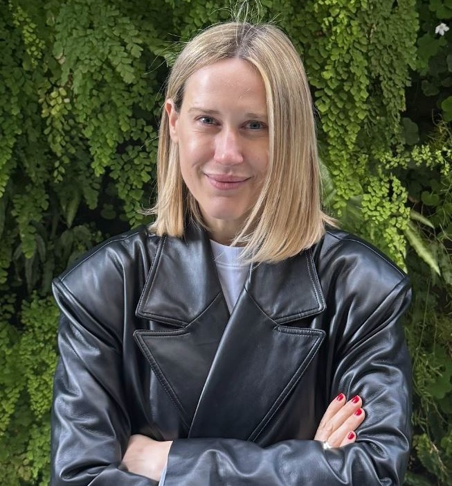 Person in a black leather jacket standing with arms crossed in front of a lush green bush.