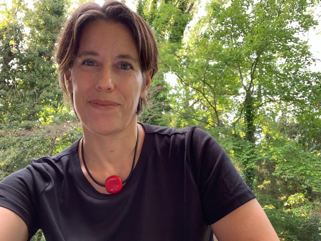 A smiling woman with chin-length hair wearing a tshirt and necklace with a large red stone