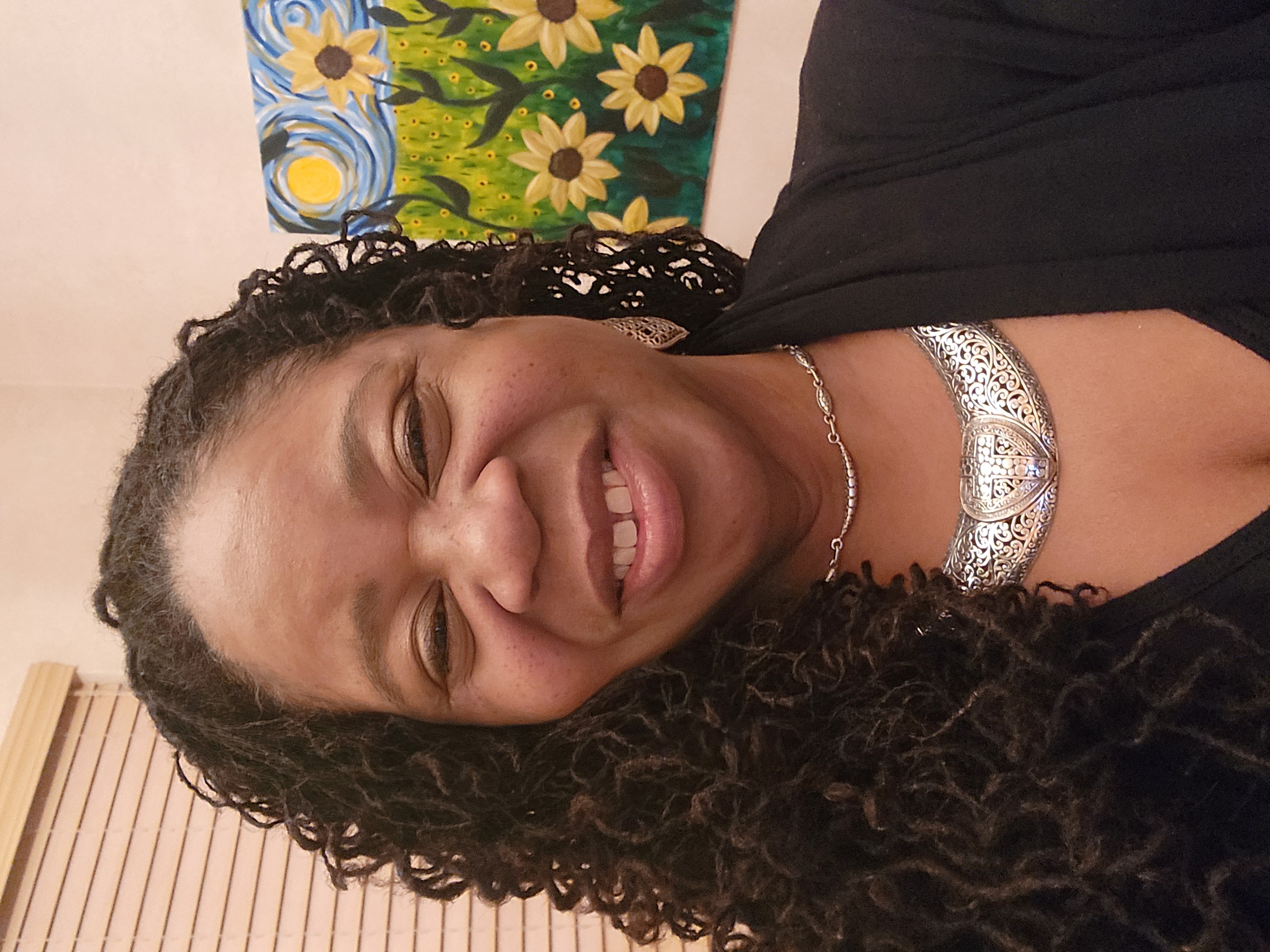 A smiling person wearing silver necklaces stands in front of a painting of sunflowers