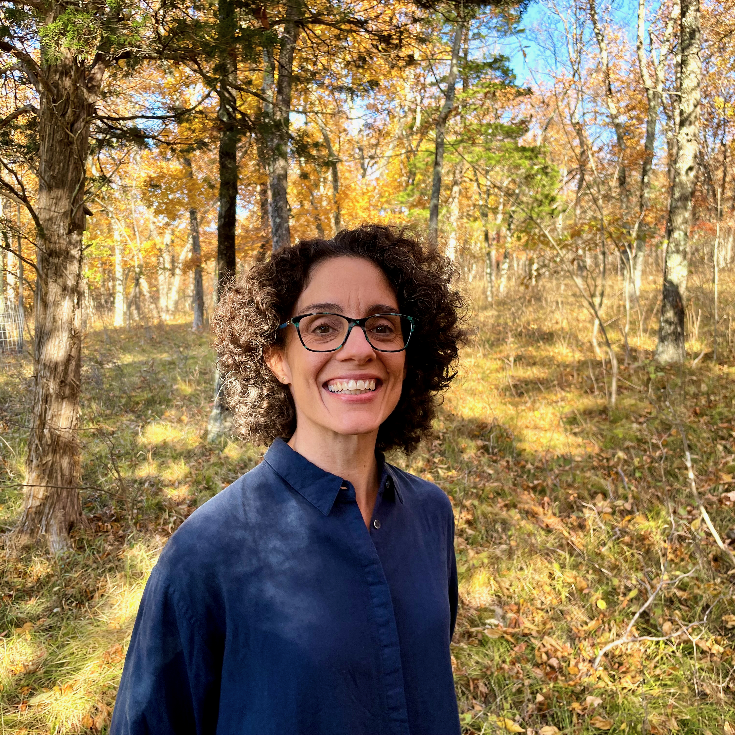 A smiling woman wearing eyeglasses stands amidst trees in a park