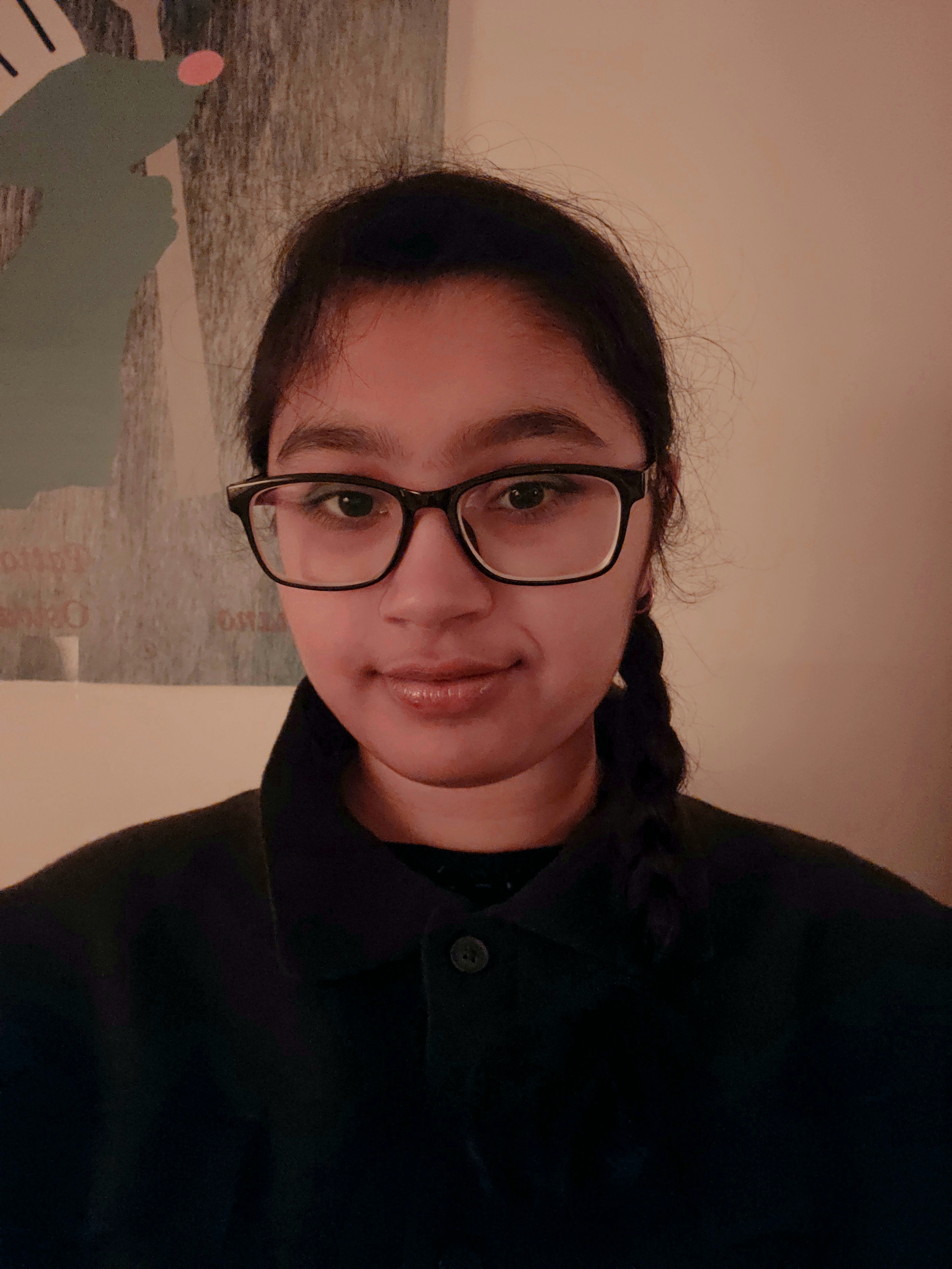 A person with glasses and a braided hairstyle smiles softly in a warmly lit room. A poster with abstract art is visible in the background.
