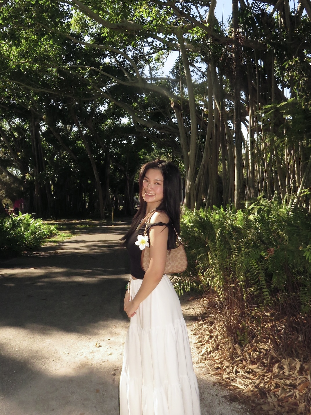 A woman smiles in a sunlit forest path lined with tall trees. She wears a black top, white skirt, and holds a plumeria flower, conveying tranquility.