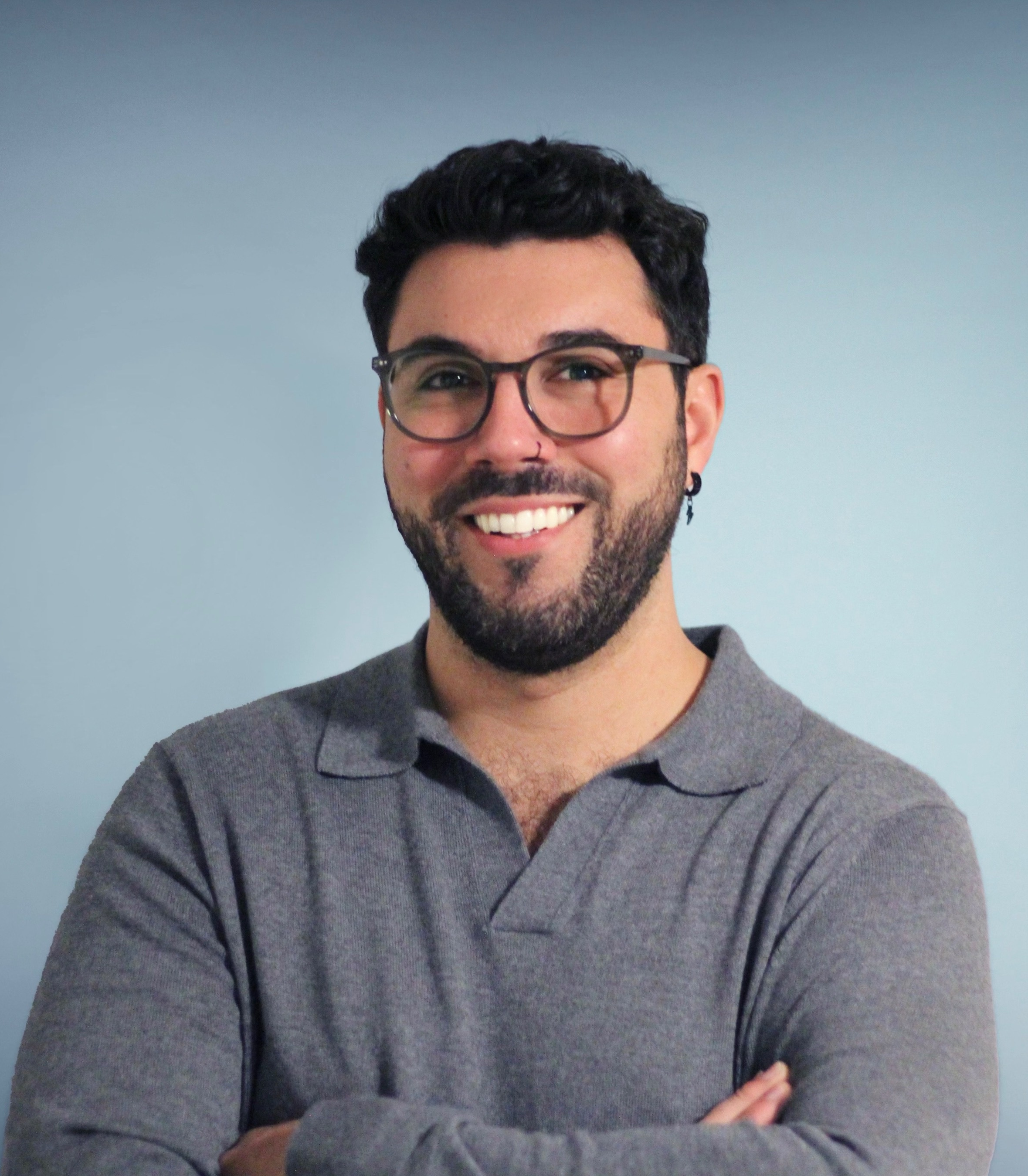 alt="A smiling man with glasses and a beard stands against a light blue background. He is wearing a gray, long-sleeved, collared shirt and has his arms crossed