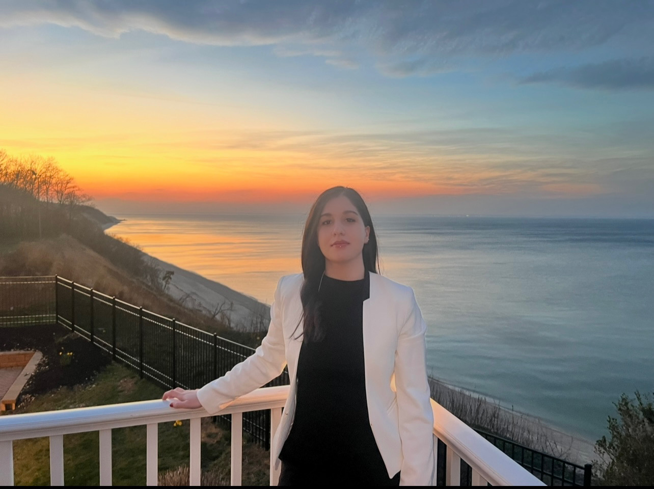 Woman in a white blazer stands on a balcony overlooking a serene ocean at sunset. The sky is vibrant with hues of orange, pink, and blue. Tranquil mood.