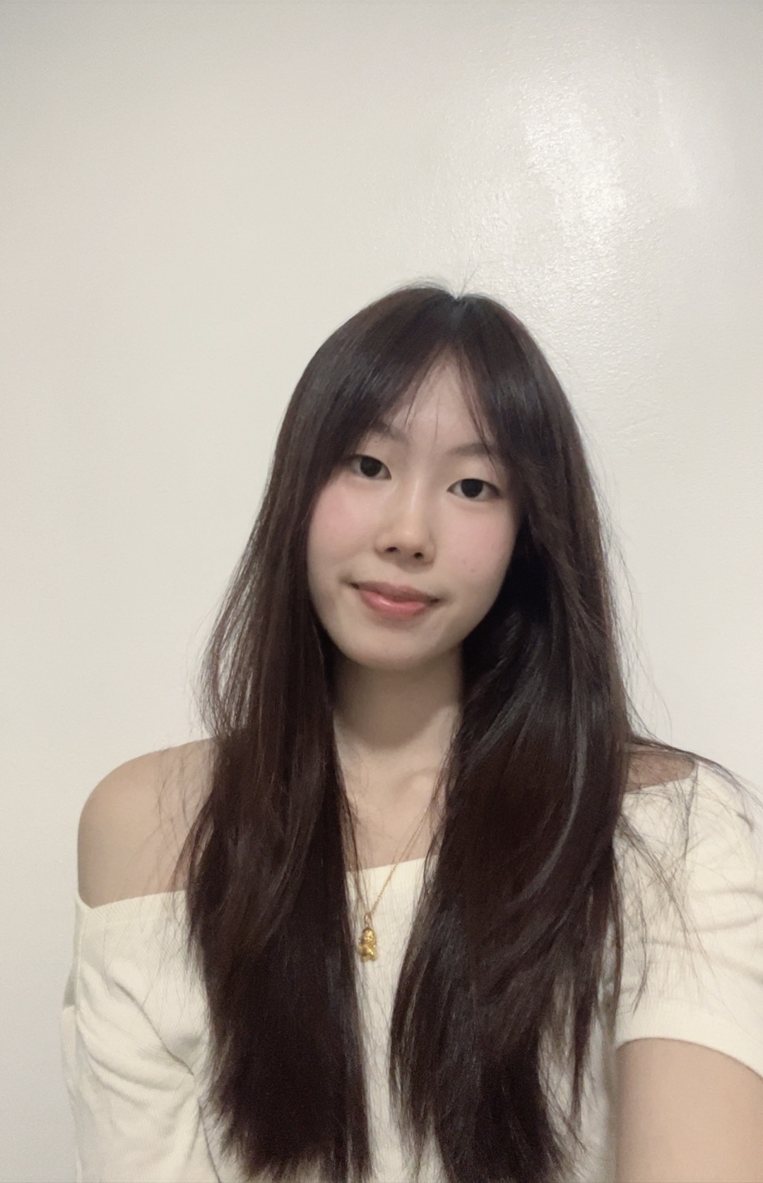 Young woman with long dark hair, wearing an off-shoulder cream top, smiles softly against a plain white background. 