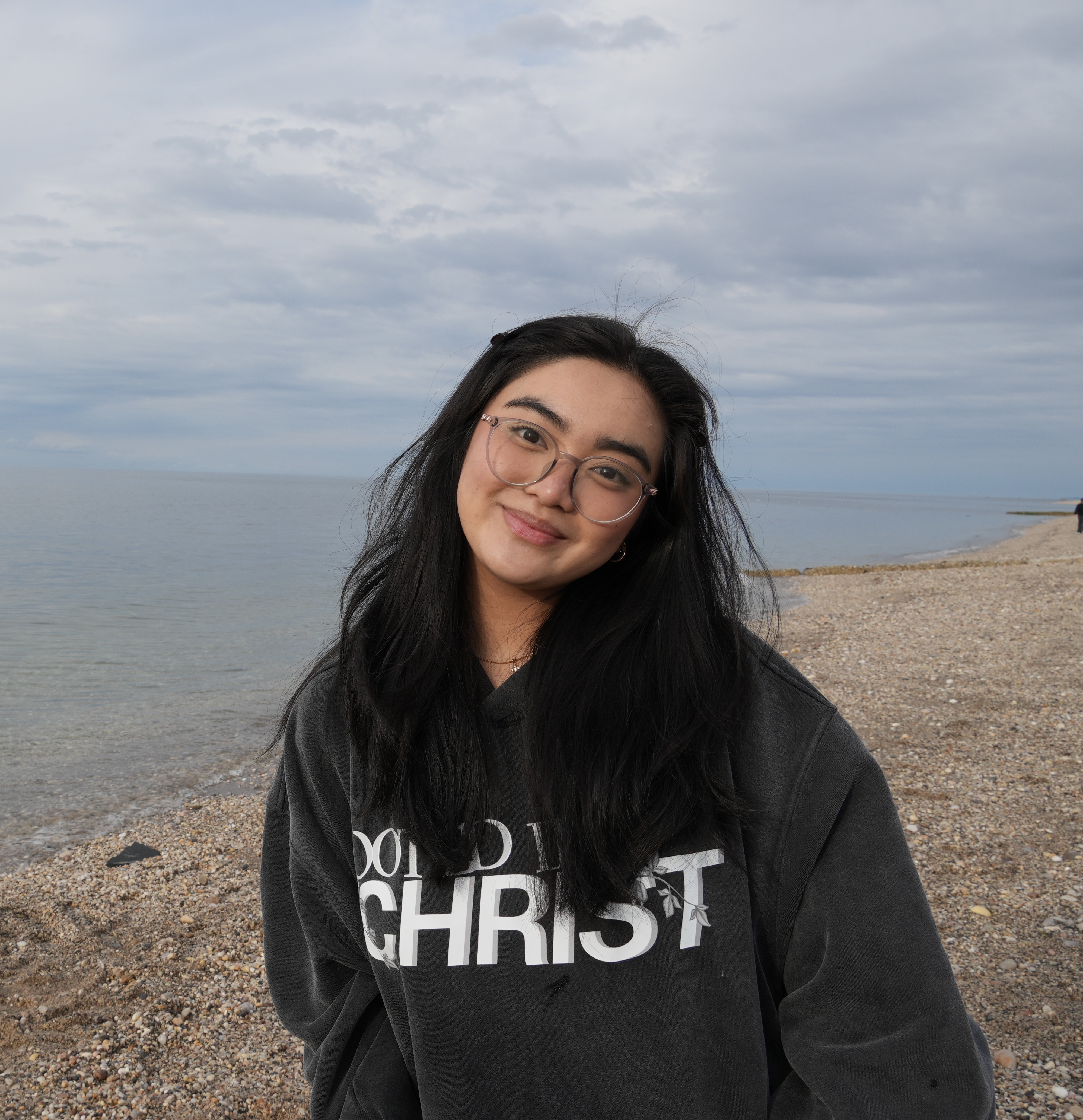 A smiling person with long dark hair and glasses stands on a pebbly beach under a cloudy sky, wearing a dark sweatshirt with white lettering.