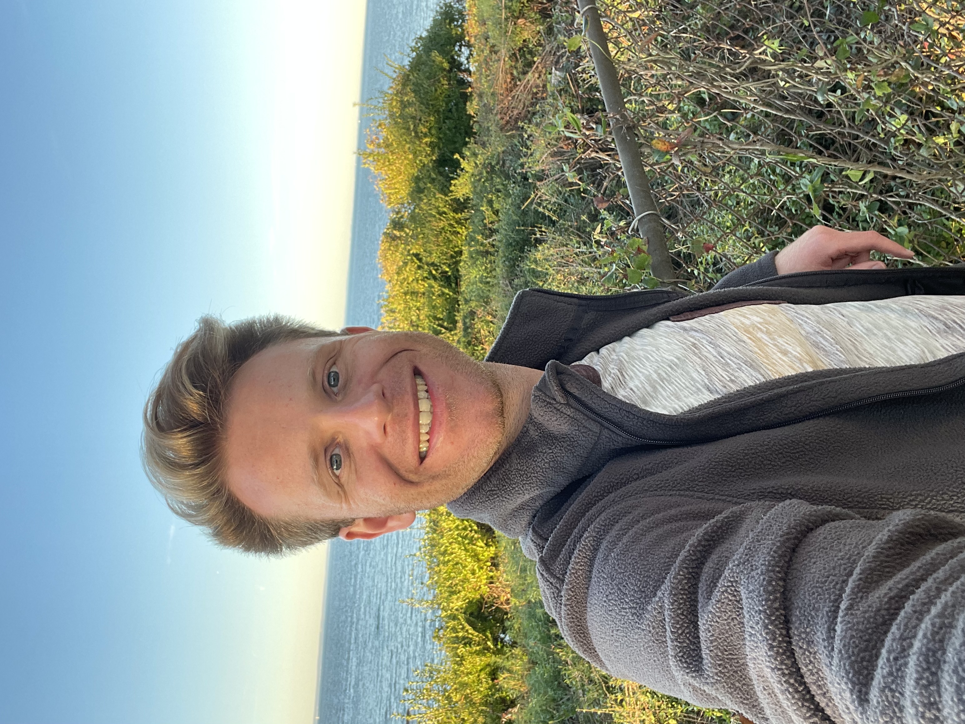 A person takes a selfie, smiling by the coast. Behind them, lush greenery and a calm sea stretch under a clear blue sky, evoking a cheerful mood.