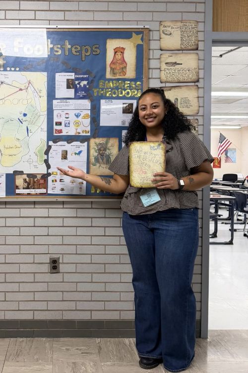 A smiling woman stands beside a bulletin board full of educational history materials, pointing at it. She holds a book, conveying a welcoming and informative atmosphere.