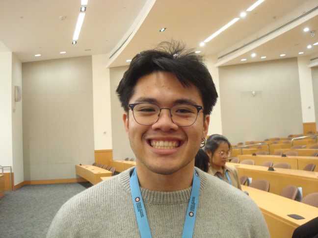 A person with glasses and a big smile stands in a lecture hall, wearing a lanyard and a gray sweater. The room is filled with empty chairs.