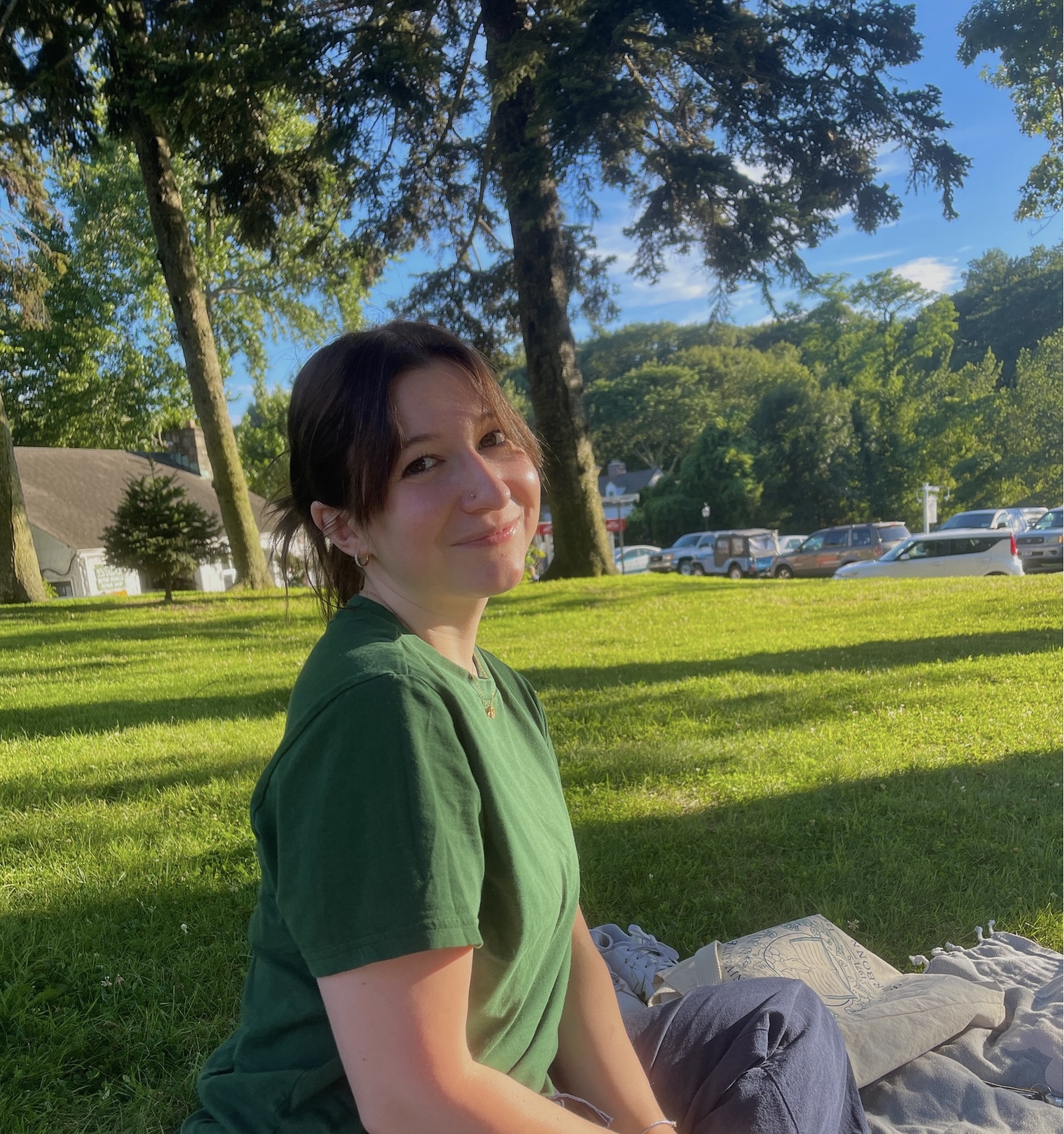 A person in a green shirt sits on the grass, smiling at the camera. Sunlight filters through trees, casting dappled shadows in a tranquil park setting.