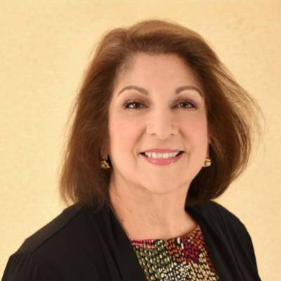 A portrait of a woman with shoulder-length brown hair and a friendly smile. She is wearing a black cardigan over a colorful, patterned top and small gold earrings. She is positioned against a warm, light-beige textured background.