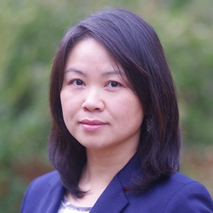 A professional headshot of a woman with shoulder-length dark hair looking toward the camera. She is wearing a dark blue blazer. The background is a soft-focus, natural green outdoor setting.