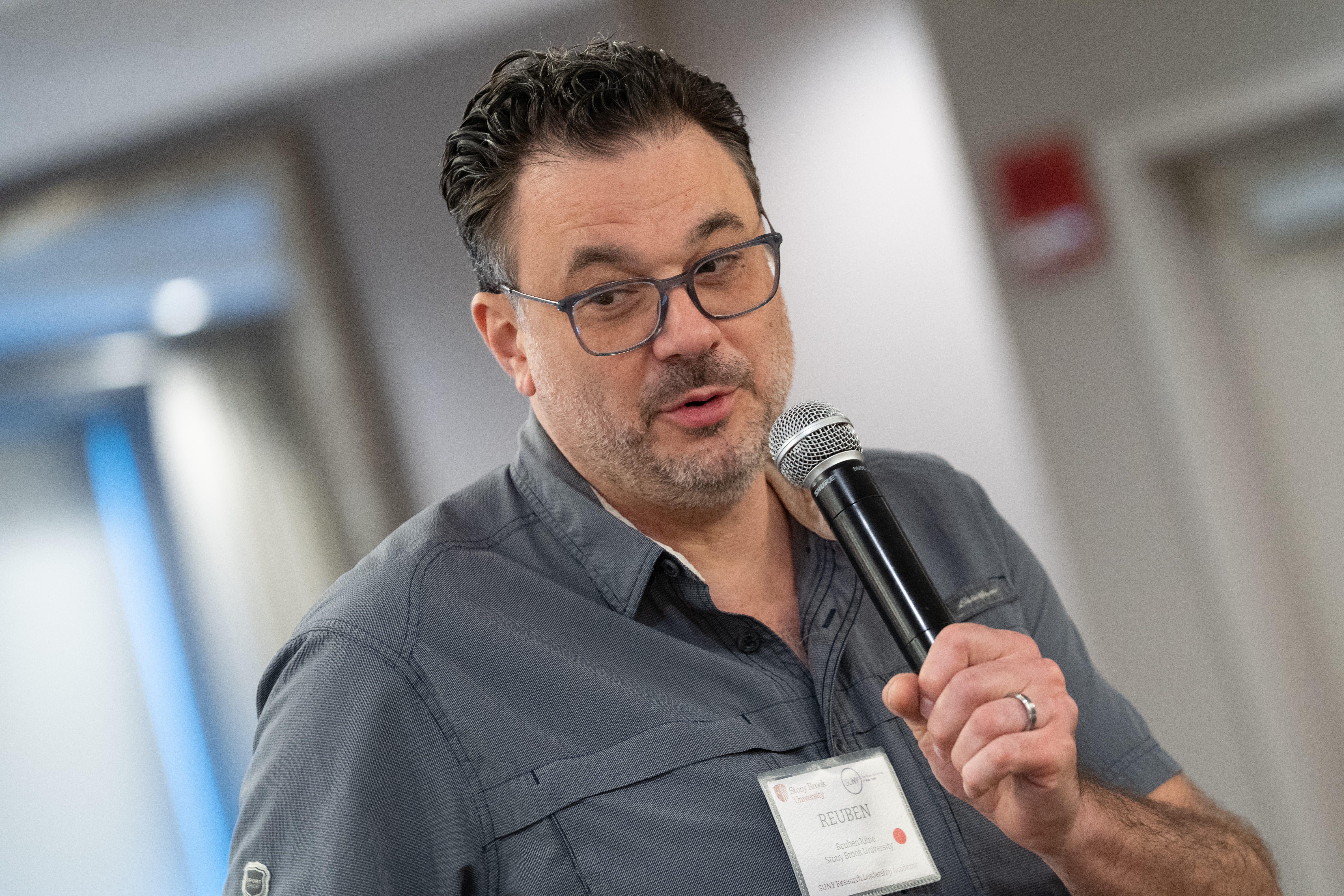 A man with dark, wavy hair and glasses, identified as "REUBEN" on his name tag, speaks into a handheld microphone. He is wearing a grey button-down shirt and a wedding band on his left hand as he looks slightly toward the side.