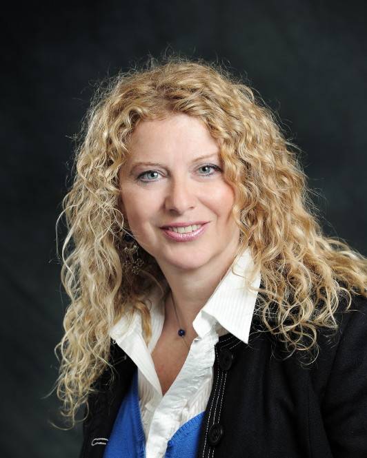 A smiling woman with long, voluminous blonde curly hair. She is wearing a black jacket over a white collared shirt and a blue sweater.