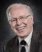 A professional headshot of an older man with white hair and glasses, smiling broadly. He is wearing a dark suit jacket over a light-colored checkered shirt and a dark necktie. The background is a solid, dark neutral tone.