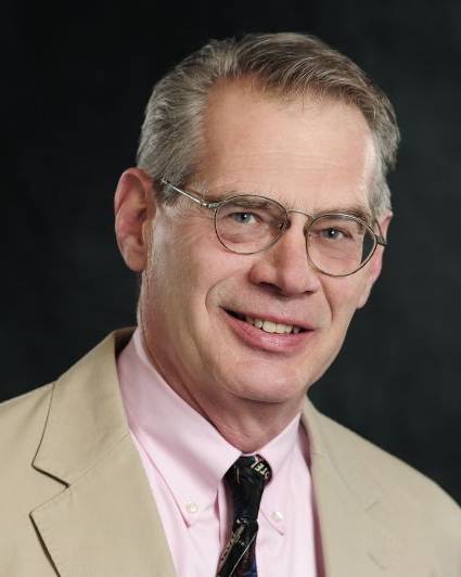 A headshot of a smiling man with short, greying hair and glasses. He is wearing a tan blazer over a light pink button-down shirt and a dark necktie with a subtle pattern. The photo is set against a solid dark background.