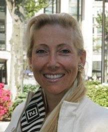 A portrait of a woman with light blonde hair pulled back, smiling at the camera. She is wearing a white jacket over a black and white geometric patterned top, standing in a bright outdoor setting with greenery and buildings in the background.