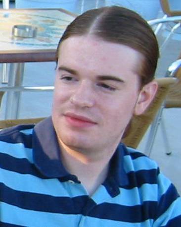 A portrait of a young man with light brown hair neatly parted in the middle and slicked back. He is looking off to the side with a neutral expression, wearing a polo shirt with horizontal navy and light blue stripes. The background is a bright, slightly blurred outdoor setting.