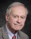 A portrait of an older man with thin gray hair and a light complexion. He is looking at the camera with a subtle, neutral smile, wearing a brown patterned blazer, a light blue dress shirt, and a red patterned tie. He is positioned against a dark, textured gray background.