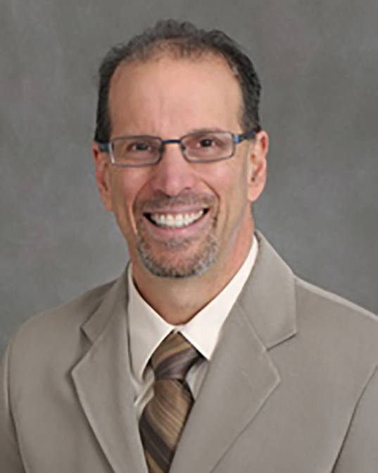 A professional headshot of a smiling man with short dark hair, glasses, and a groomed goatee. He is wearing a tan suit jacket, a white dress shirt, and a brown striped necktie. The photo is set against a plain, light-grey studio background.