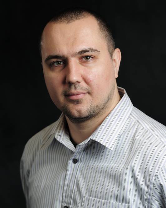 A man with a short buzz cut and a slight beard, looking directly at the camera with a neutral expression. He is wearing a light-colored, thin-striped button-down shirt.
