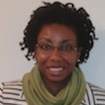 A professional headshot of a woman with short, curly dark hair and glasses, smiling. She is wearing a black-and-white striped blazer paired with a light green scarf. The background is a plain, neutral white.