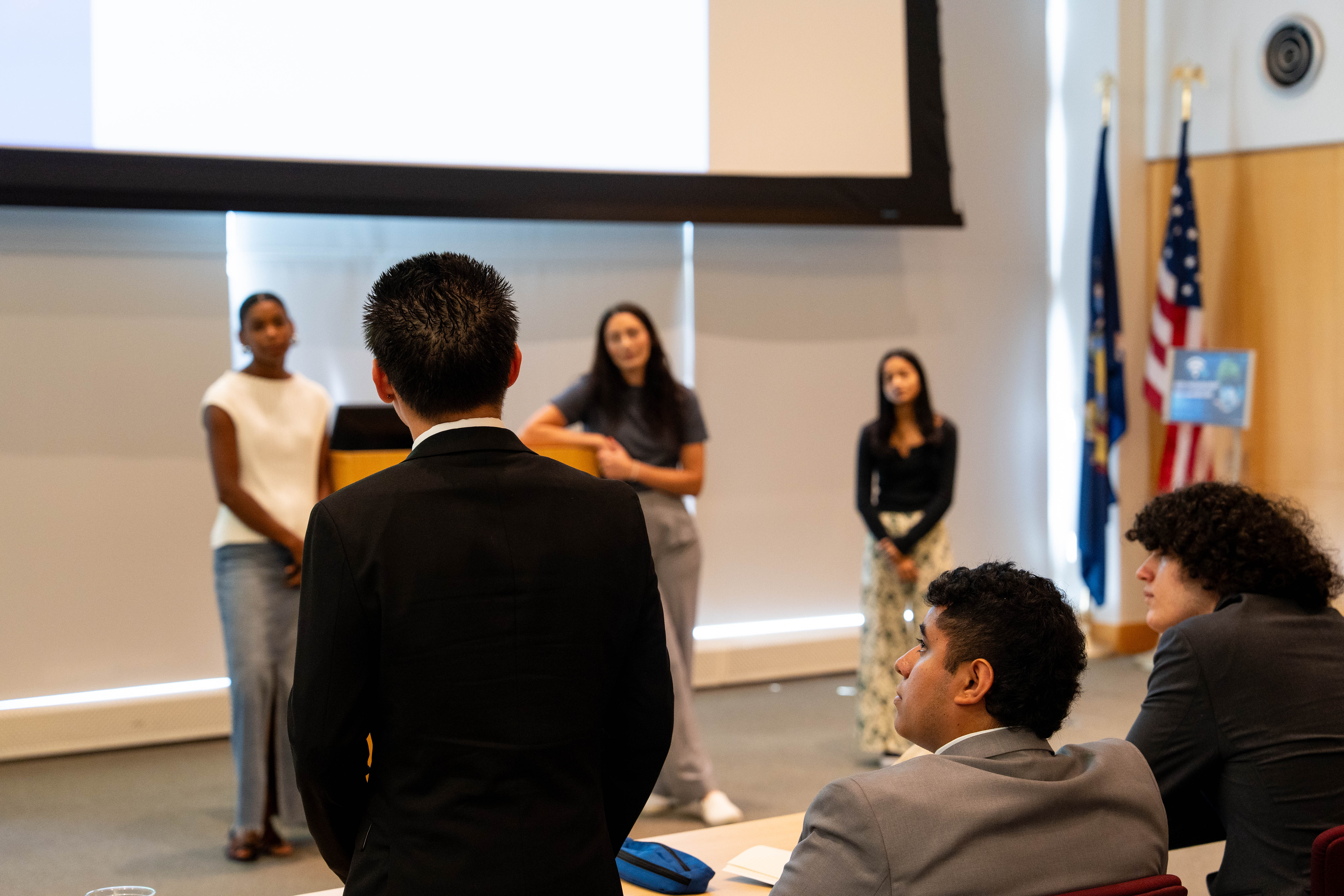 A man in a black suit stands with his back to the camera, facing three women who are standing at the front of a room, possibly during a presentation or Q&A session.  The three women are standing in front of a large, blank projection screen; one wears a white sleeveless top and long denim skirt, the middle wears a grey t-shirt and light trousers, and the one on the right wears a black long-sleeved top and patterned pants.  In the foreground, two other men are seated at a table, looking up toward the presenters.  The background includes wood-paneled walls, an American flag, and a New York State flag.