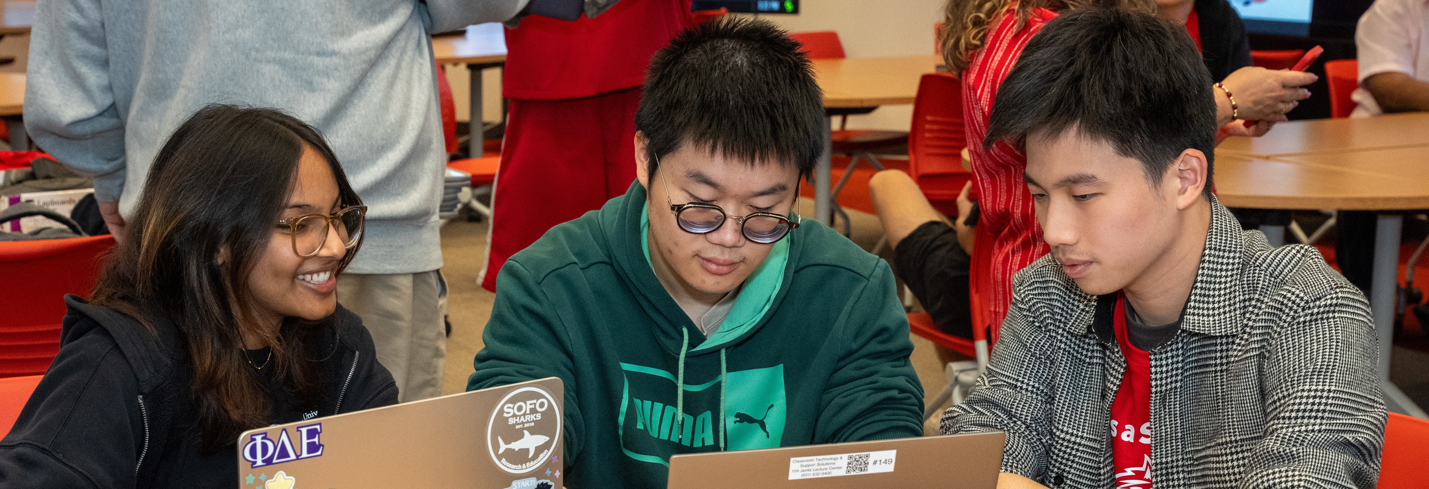 Three students—a young woman and two young men—sit together at a table, focused on a laptop. They are in a well-lit indoor space with red chairs and other people in the background. The student on the left wears glasses and is smiling, while the others look on with concentration. The laptop in the foreground features several stickers, including Greek letters and a shark logo.