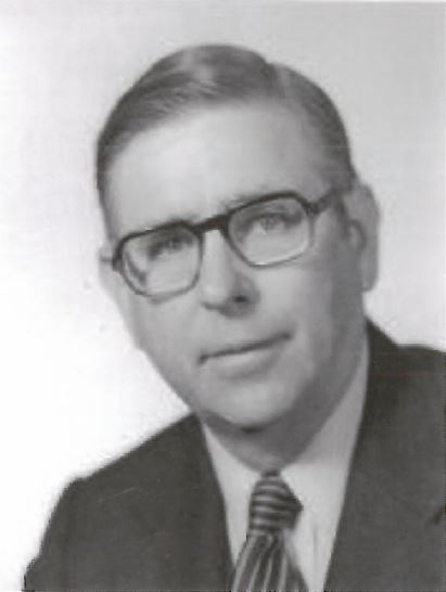 A black-and-white vintage portrait of a man with light-colored hair and glasses. He is wearing a dark suit jacket, a light-colored dress shirt, and a diagonally striped tie. He is looking toward the camera with a neutral expression. The background is a plain, light-colored gradient.