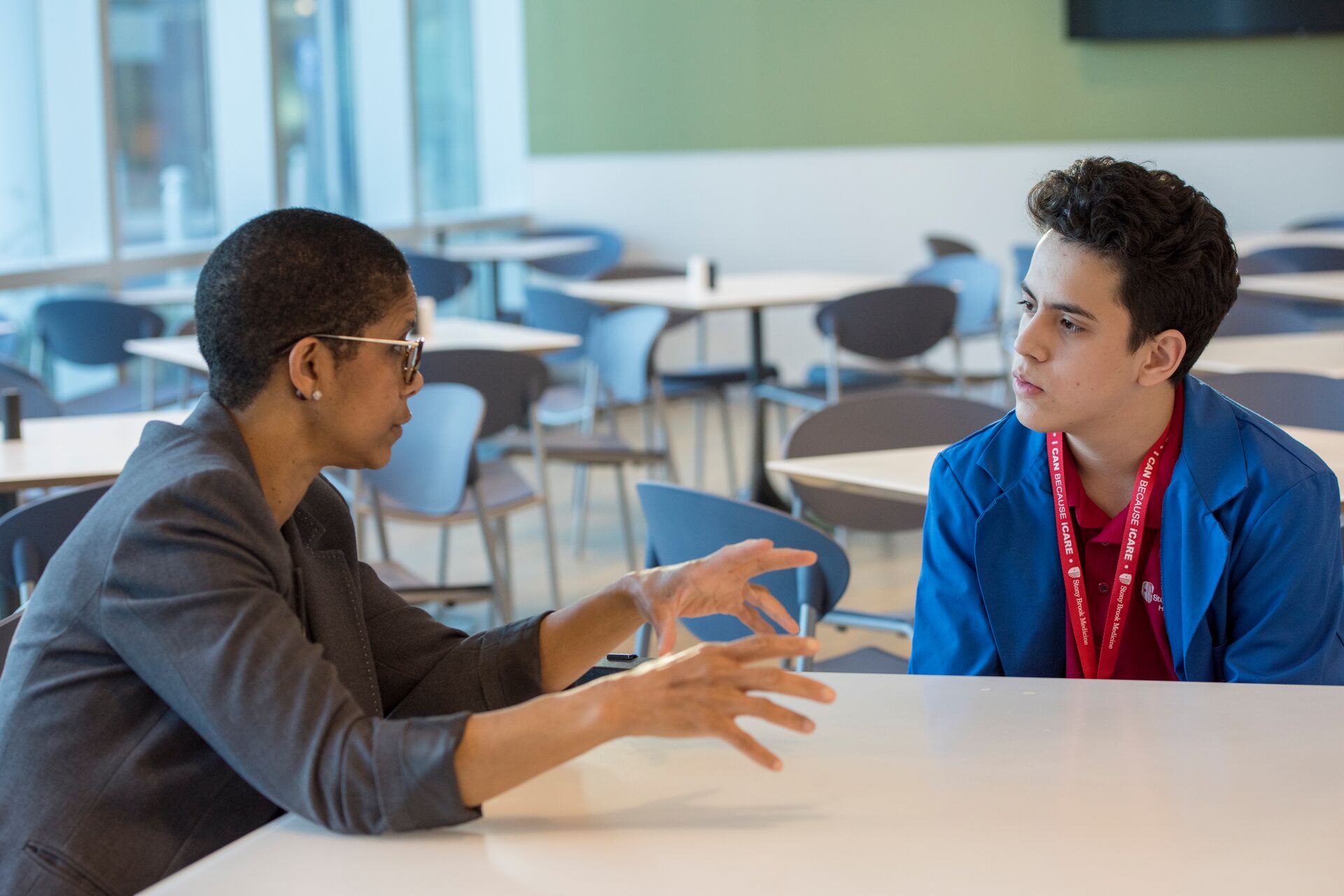 A woman with short-cropped dark hair and glasses sits at a white table in a bright, modern cafeteria or lounge area, gesturing with both hands while speaking to a young man. The woman is wearing a dark grey blazer. The young man, listening intently, has dark, wavy hair and is wearing a blue jacket over a red collared shirt with a red "I CAN BECAUSE ICARE" lanyard from Stony Brook Medicine. The background is filled with numerous empty white tables and blue chairs, with large windows on the left.