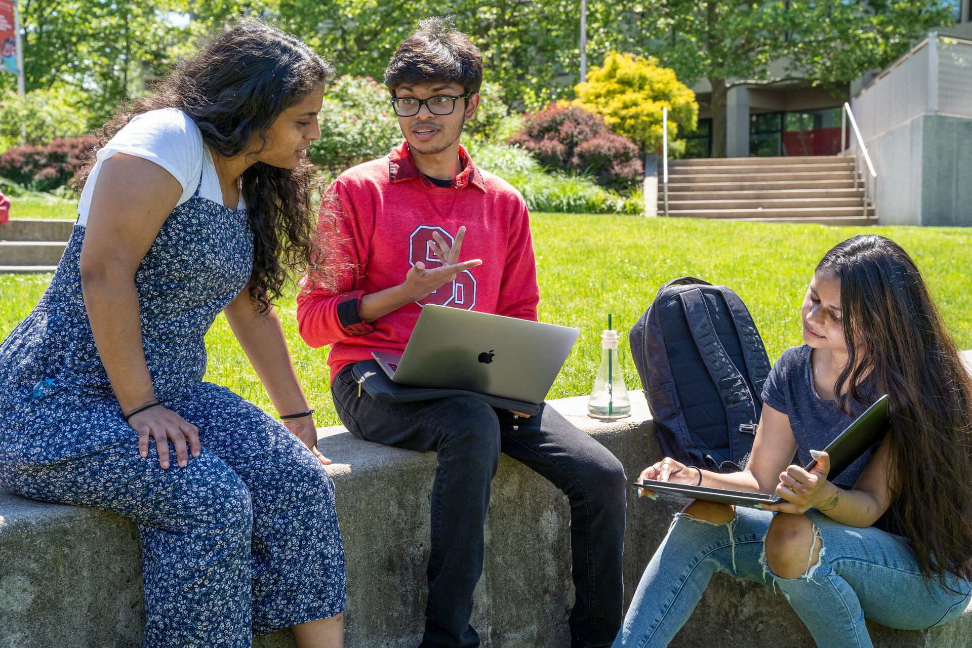 Three students are sitting outdoors on a stone ledge in a grassy campus area on a sunny day.  The student on the left is a young woman with long dark curly hair, wearing a white t-shirt under a blue and white floral jumpsuit. She is leaning toward the center, looking at a laptop screen.  The student in the middle is a young man with glasses and short dark hair, wearing a red sweatshirt with an "S" logo. He is gesturing with one hand while holding an open silver laptop on his lap.  The student on the right is a young woman with long dark hair, wearing a dark blue t-shirt and ripped light-wash jeans. She is looking down at a tablet or notebook in her lap.  A dark backpack and a clear water bottle sit on the ledge between the students. In the background, there is a green lawn, lush trees, and a set of concrete stairs leading up to a campus building.