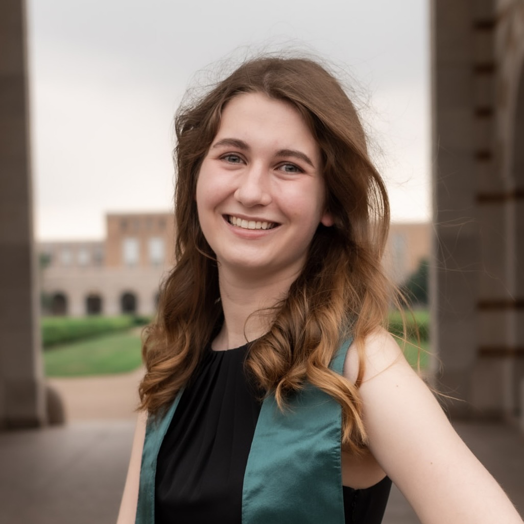 A professional headshot of a smiling young woman with long, wavy brown hair, wearing a black top and a green graduation-style stole. She is standing outdoors in a courtyard, with large stone pillars framing her and a college campus building visible in the soft-focus background.