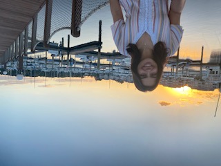A young woman with shoulder-length dark hair, wearing a white and yellow striped V-neck top, stands on a wooden pier at sunset. Behind her, calm water reflects the orange sky, and several white boats are docked in a marina against a backdrop of buildings.