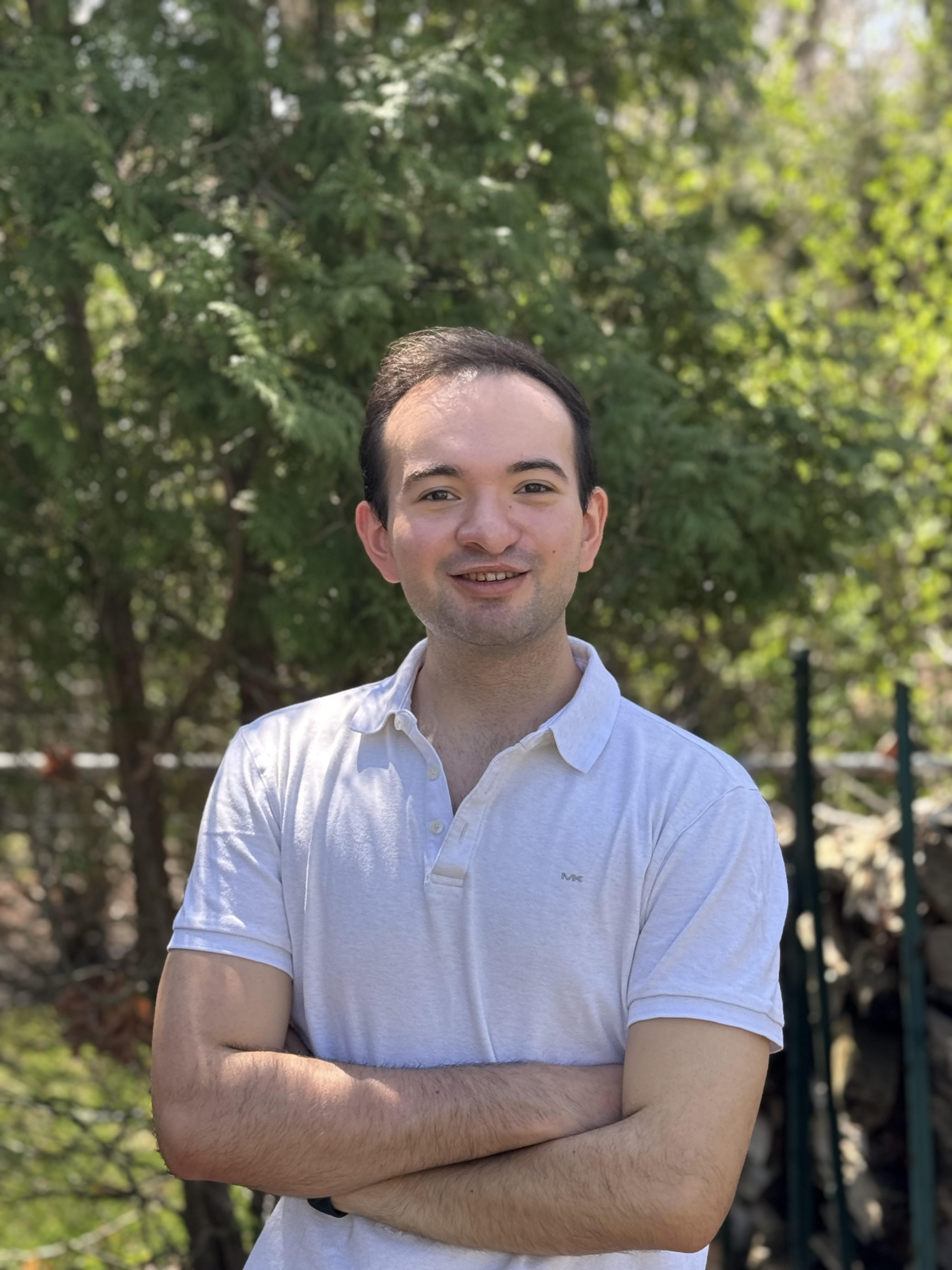 Johan Arias Chacon A portrait of a young man with short dark hair, smiling and posing with his arms crossed. He is wearing a white short-sleeved polo shirt. The photo is taken outdoors in a backyard setting, with a large evergreen tree and other green foliage providing a natural, soft-focus background.