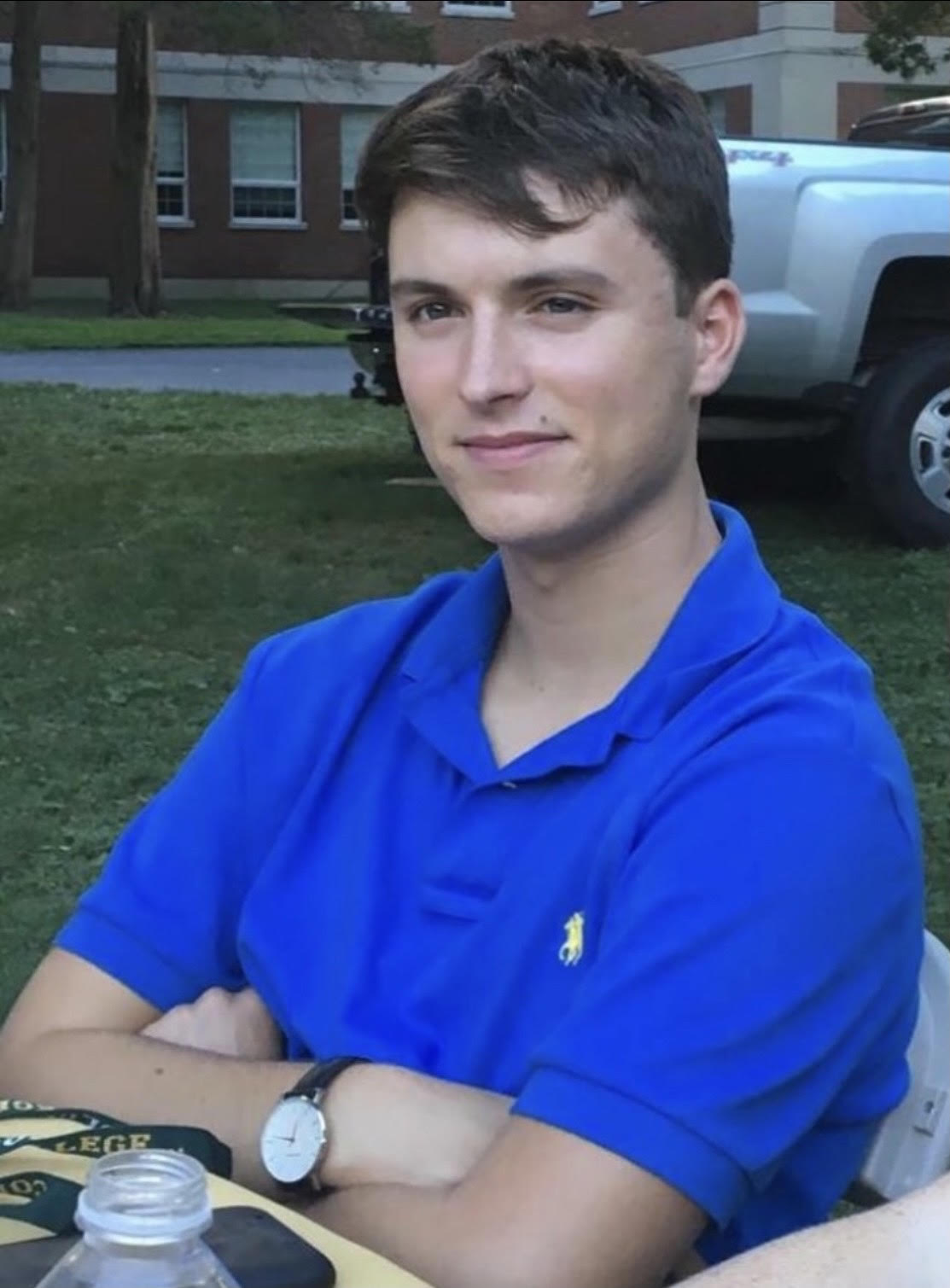 A young man with short dark hair, wearing a bright blue polo shirt with a yellow logo, sits at an outdoor table with his arms crossed. He is looking slightly off-camera with a neutral expression. In the background, there is a grassy area, a brick building, and a vehicle.