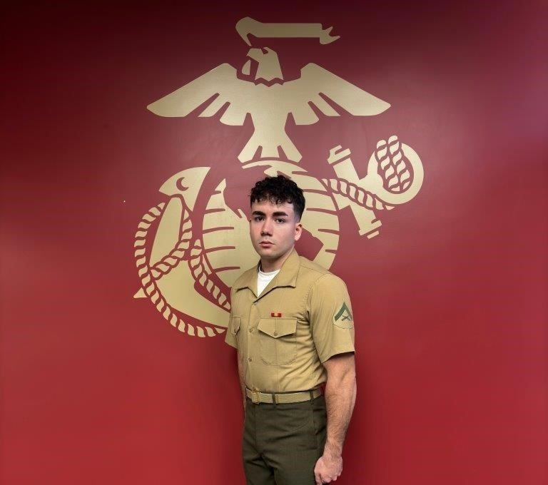 A portrait of a young man, Noah Kirchner, in a United States Marine Corps Service C uniform. He is standing at attention against a solid red wall that features a large gold Eagle, Globe, and Anchor emblem. He wears a short-sleeved khaki button-down shirt with a marksmanship badge above the left pocket and a rank insignia on the sleeve, paired with olive green trousers and a web belt.