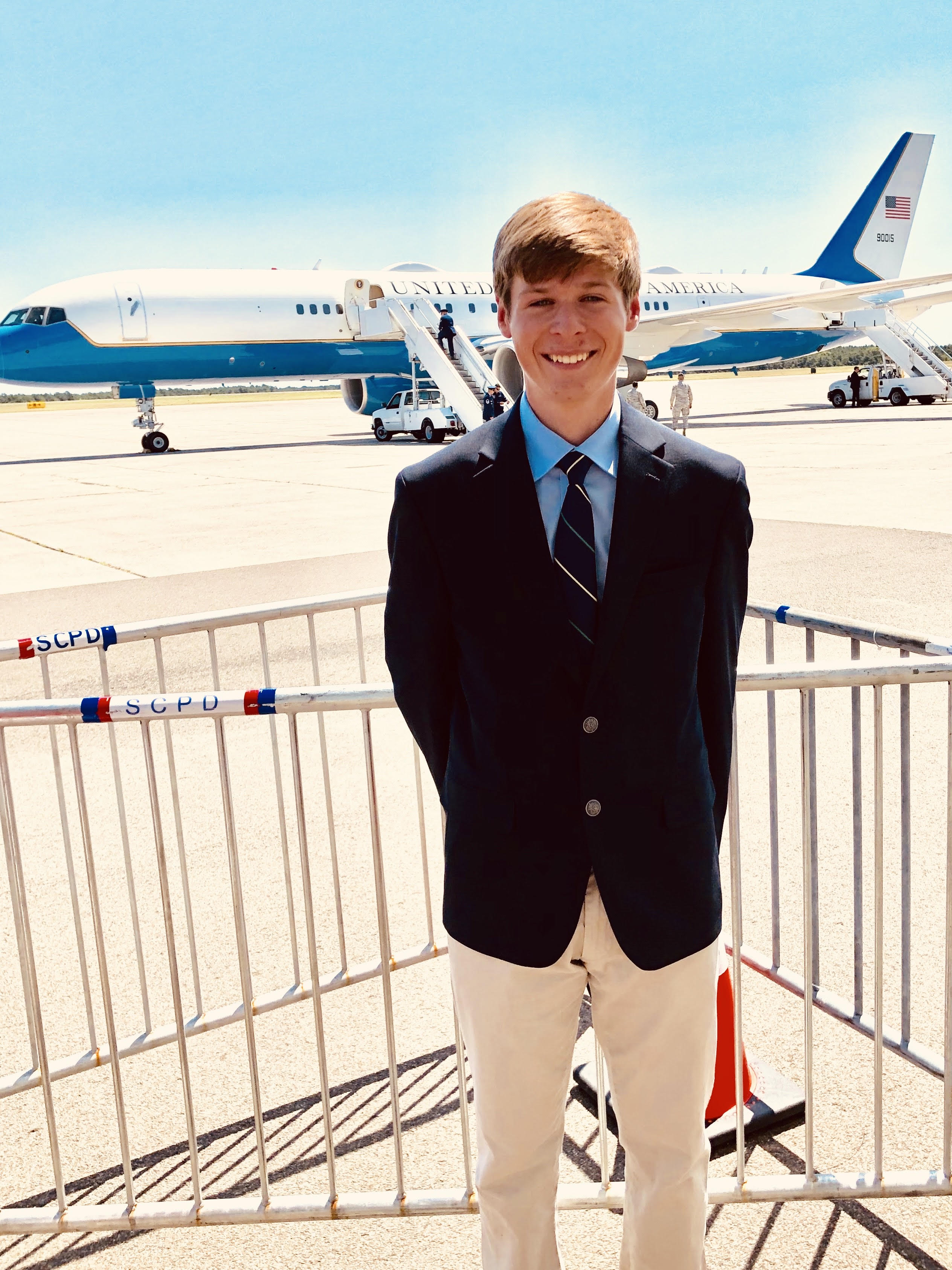 A professional headshot of a smiling young man with short blonde hair, wearing a dark blazer, a light blue collared shirt, and a striped tie. He is standing outdoors behind a metal security railing on an airfield. In the background, a large "United States of America" aircraft (Air Force Two) is parked on the tarmac under a clear blue sky.