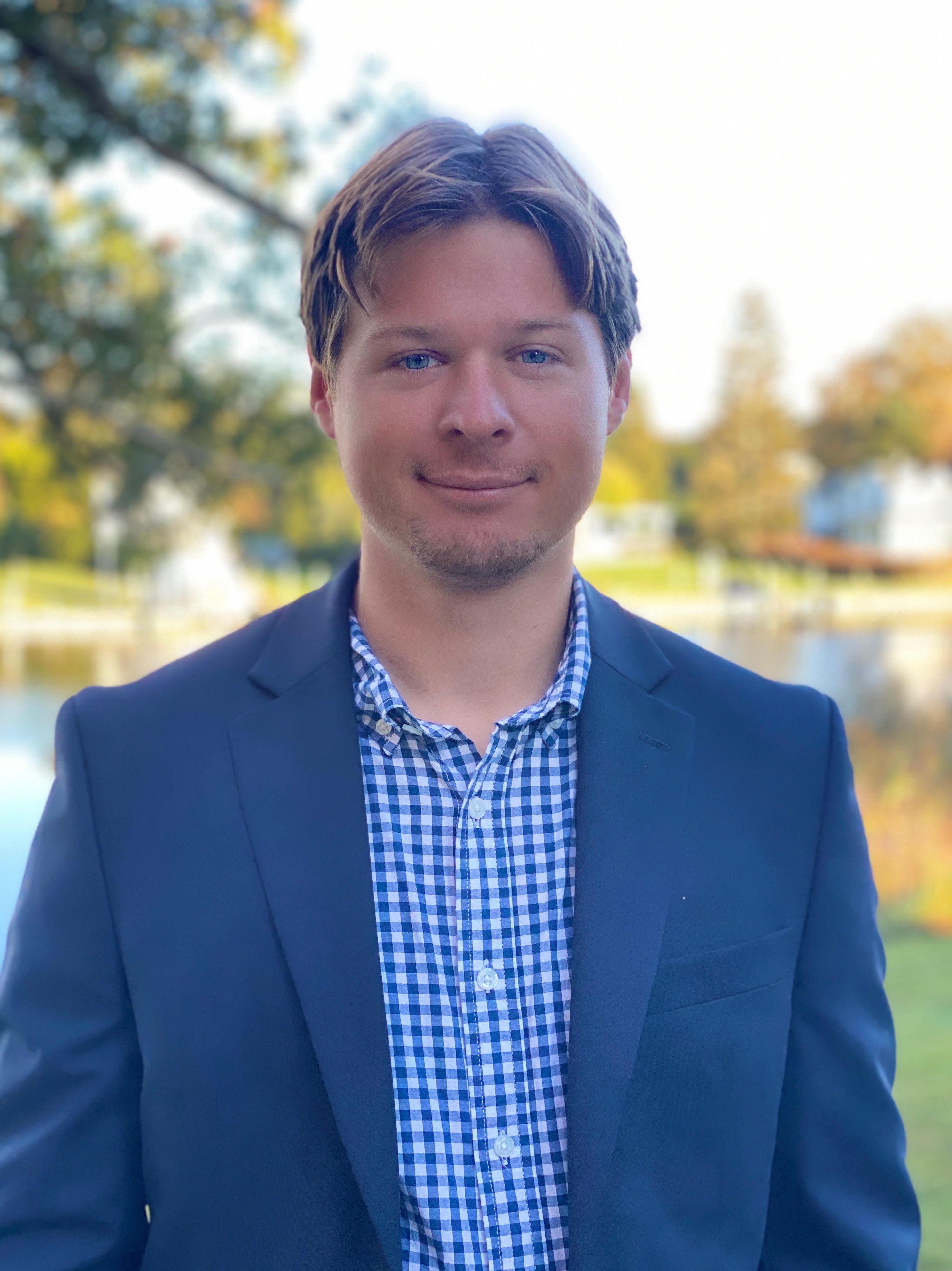 A professional headshot of a smiling young man with light brown hair and blue eyes. He is wearing a navy blue blazer over a blue and white checkered button-down shirt. The background is an outdoor setting featuring a body of water and trees in soft focus.