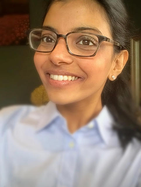 A close-up headshot of a smiling woman with dark hair pulled back, wearing tortoiseshell-patterned glasses and a light blue collared shirt. She is also wearing small, sparkling stud earrings.