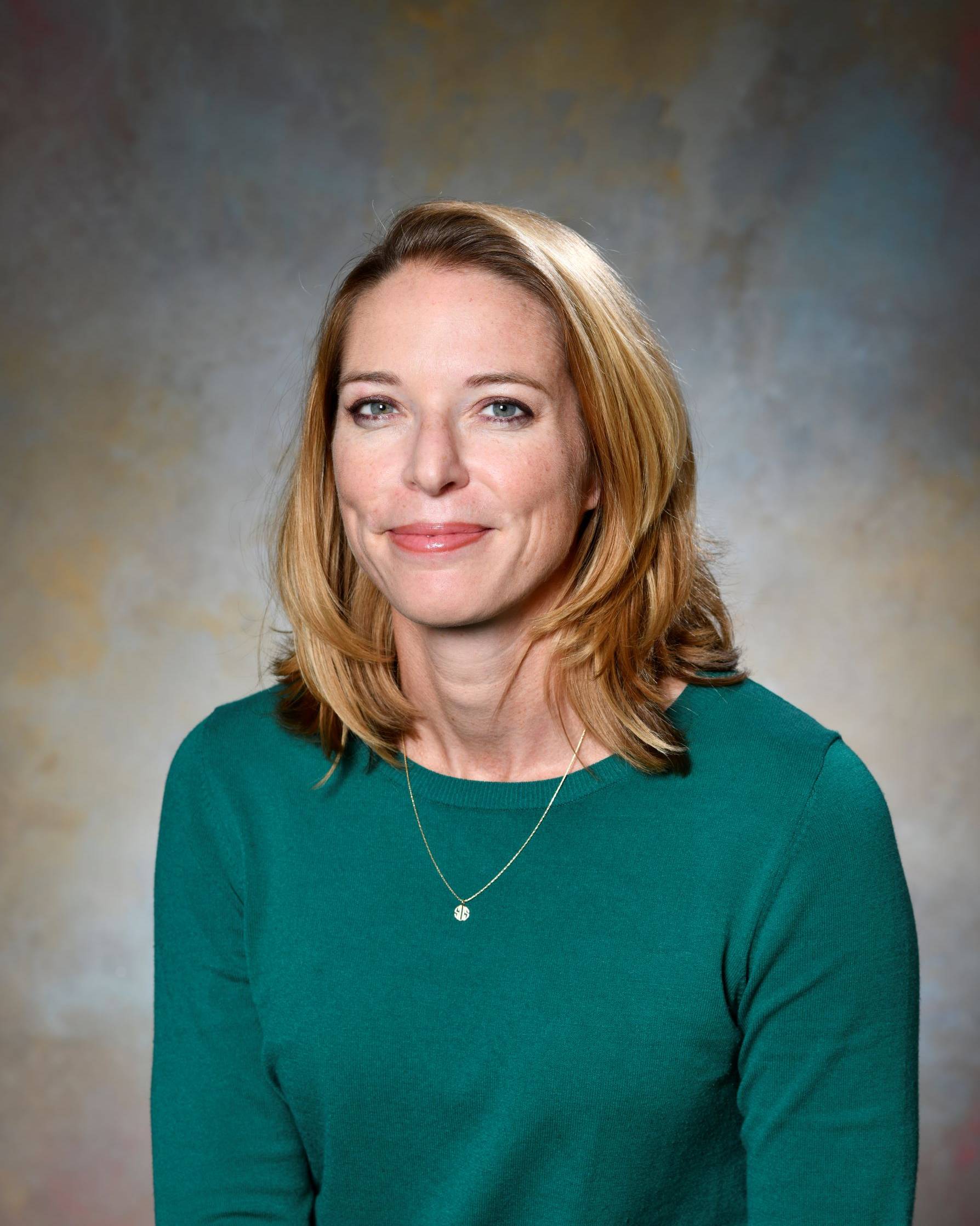 A professional headshot of a woman with shoulder-length blonde hair and blue eyes, smiling slightly at the camera. She is wearing a dark teal crewneck sweater and a delicate gold necklace with a small pendant. The background is a mottled, neutral-toned studio backdrop.