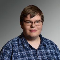 A professional headshot of a person with short brown hair and glasses, looking at the camera with a neutral expression. They are wearing a blue, black, and white checkered button-down shirt. The photo is set against a simple, solid grey background.