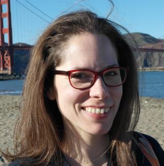 A smiling woman with long brown hair and red-framed glasses stands on a beach. In the background, the Golden Gate Bridge is visible under a clear blue sky.