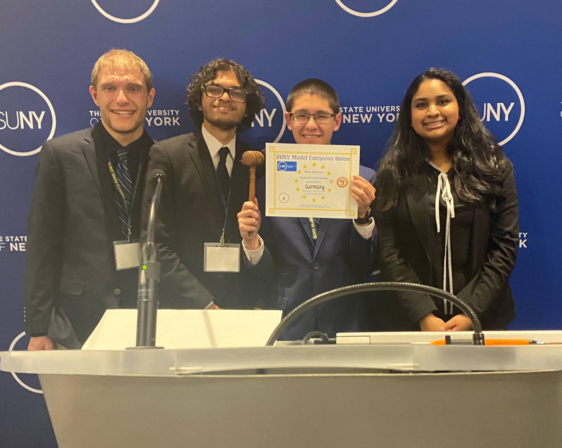 Four students—three men and one woman—stand behind a white podium in front of a blue backdrop featuring the SUNY (State University of New York) logo. The student second from the left holds a wooden gavel, and the student third from the left holds a yellow certificate that reads "SUNY Model European Union." All four are dressed in professional attire, including suits and blazers.