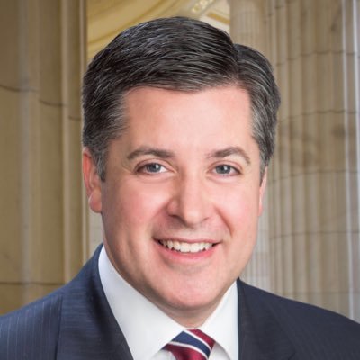 A professional headshot of a smiling man with short, dark hair. He is wearing a navy blue pinstriped suit, a white collared shirt, and a red, white, and blue striped tie. The background features light-colored, classical architectural columns.